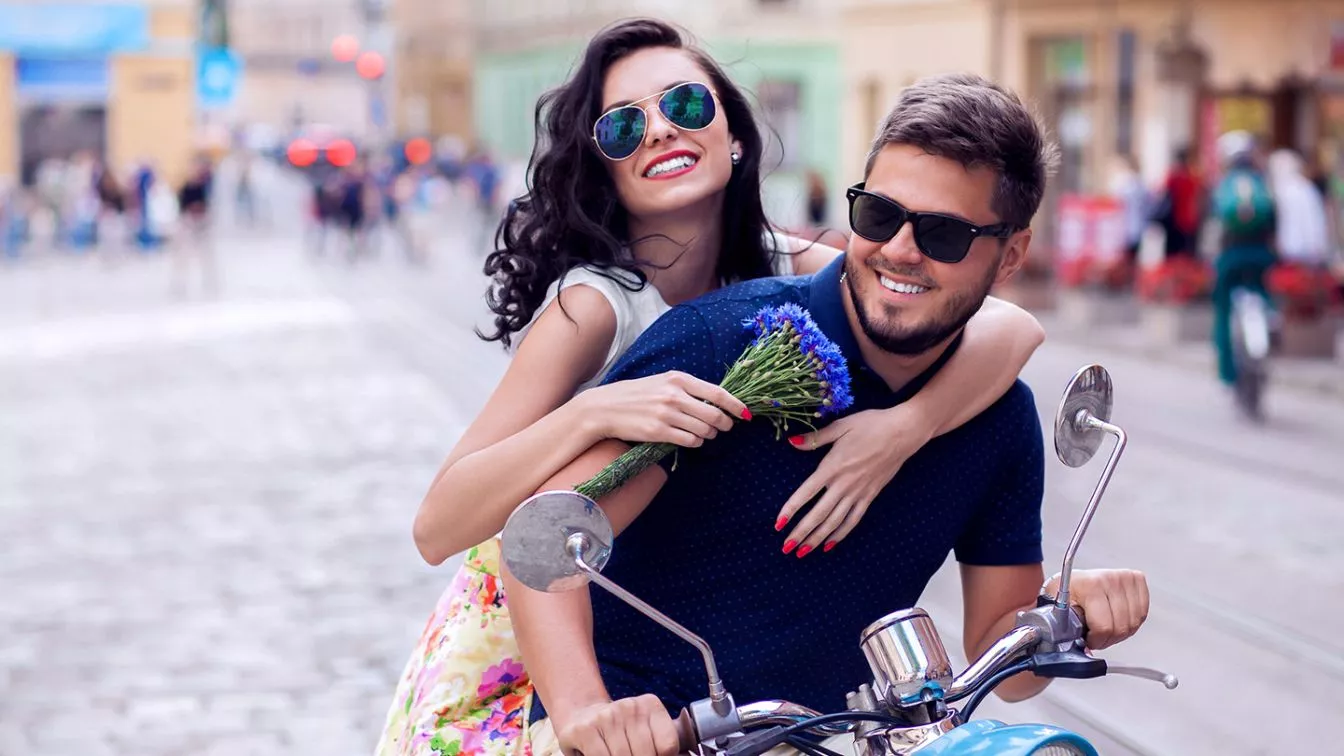 A happy couple riding a motorbike through a cobblestone street.